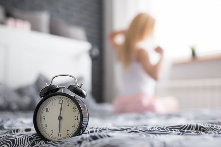 A photo of alarm clock on the bed linen and a girl in the background, who's stretching out and getting up from bed. InnerVisionPRO via Getty Images