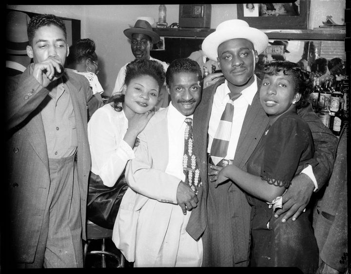 Women and men posed with arms around each other, including Erroll Garner wearing light colored suit in center, in bar or club with mirror, c. 1946
