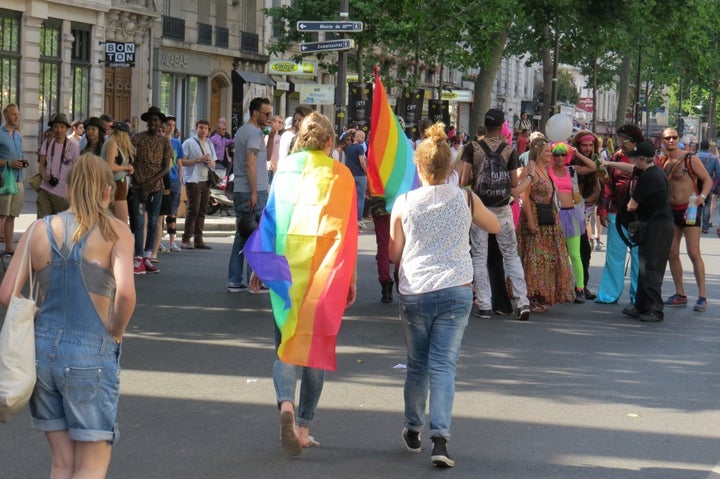 Two people face away from the camera and walk towards a larger group. The person on the left is draped in a rainbow flag.