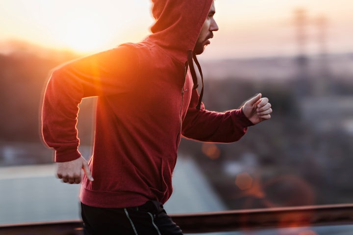 A picture of a man running and exercising beneath a clear sky, along a steal fence. He is wearing a red sweater with his hoodie pulled up and black shorts. In the distance below is some buildings and the sun is setting giving a nice warm light. Geber86 via Getty Images