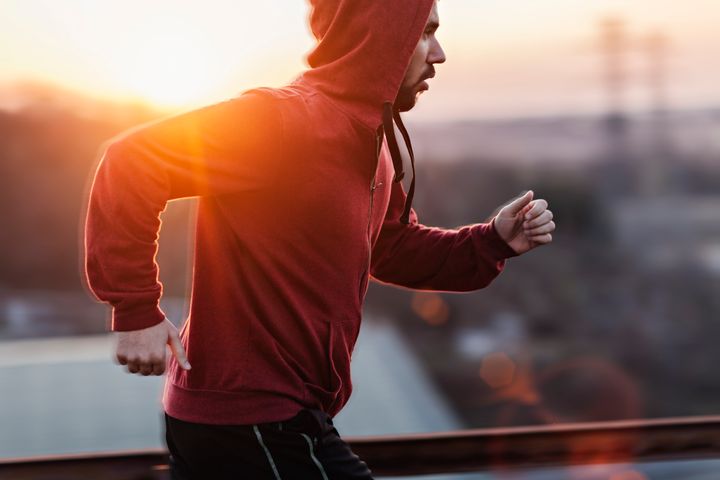 A picture of a man running and exercising beneath a clear sky, along a steal fence. He is wearing a red sweater with his hoodie pulled up and black shorts. In the distance below is some buildings and the sun is setting giving a nice warm light. Geber86 via Getty Images