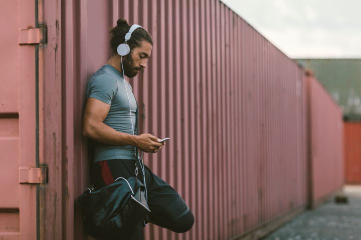 Athletic Man Resting After Running. He is running in the city every day, preparing himself for marathon race. Standing, listening music and holding his smart phone. Also texting or typing on his smart phone. Wearing gym bag. vgajic via Getty Images