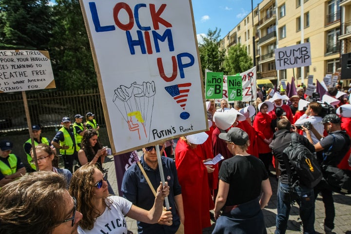 A woman holds a sign that says "Lock him up!" next to protesters dressed as handmaids.