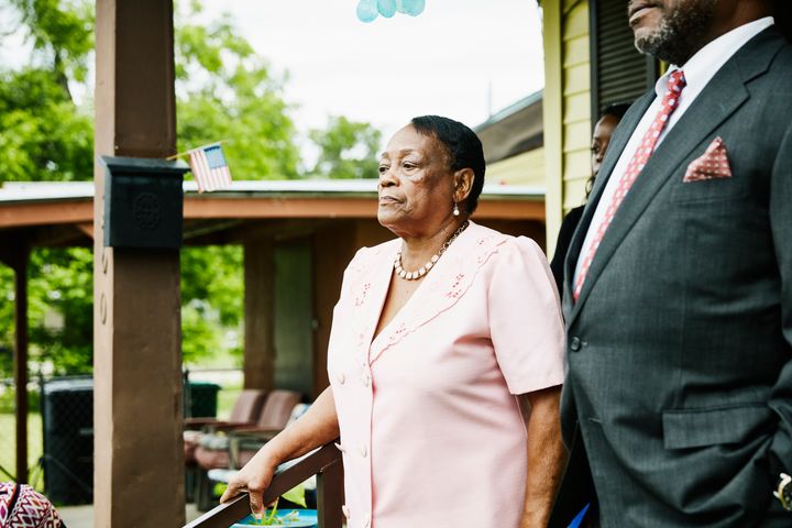 An elderly black woman stands in front of porch dressed ready for church.
