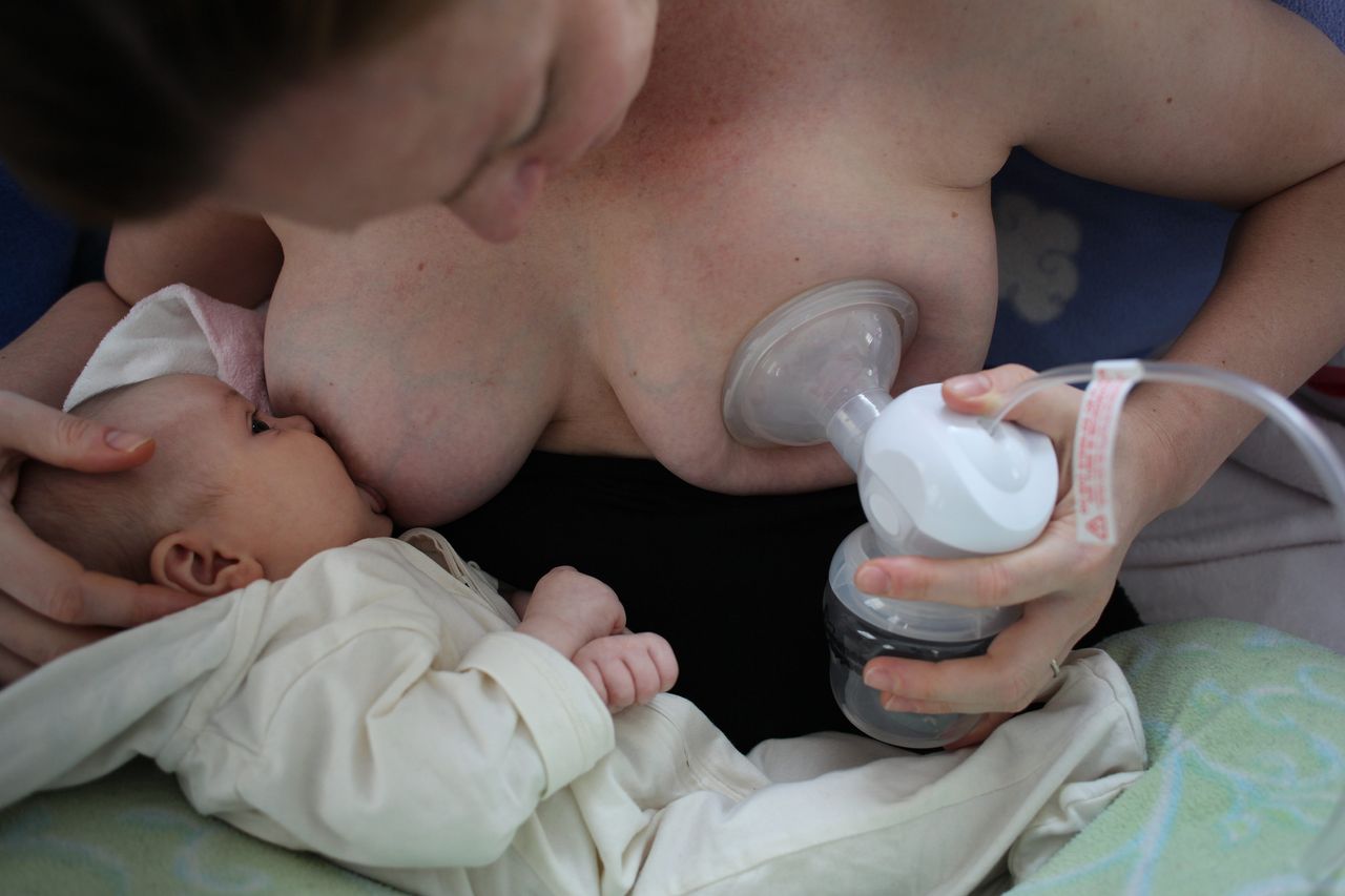 A 2-month-old baby girl baby breastfeeding as her mother uses a milk pump to express milk into a container.