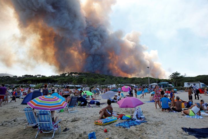 Tourists stand on the beach and watch as smoke fills the sky above a burning hillside in Bormes-les-Mimosas, in France last month