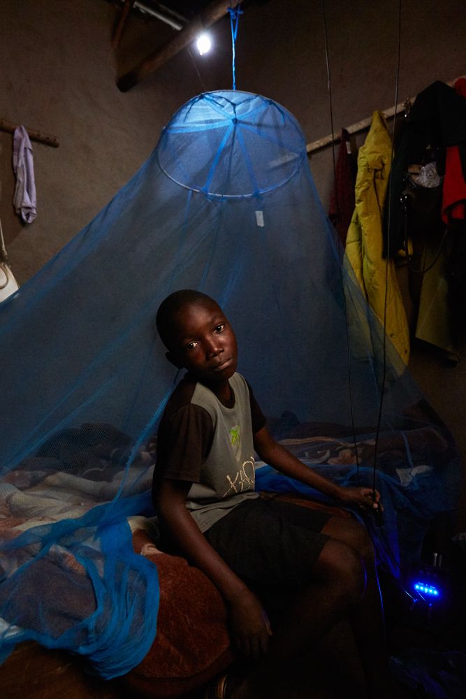 Ben Chumba (10) sits on his bed, lit by a light bulb which is powered by a solar battery, visible in the bottom right hand corner of the image, Chuita village, southern Malawi, 2017. 