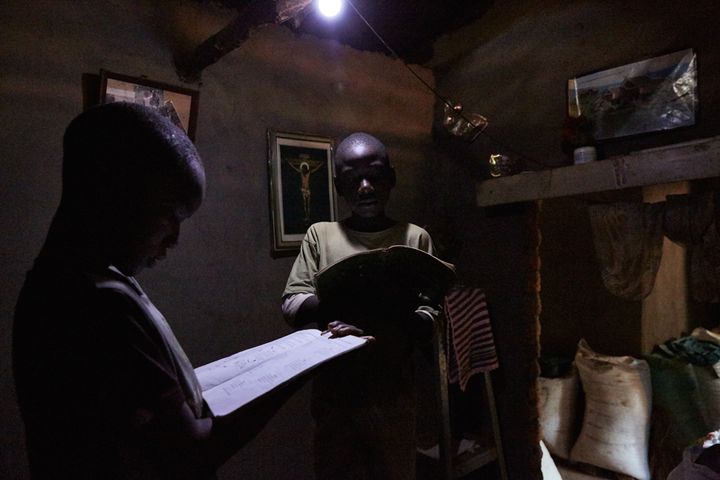 Jan Chumba (12) and his brother Ben (10) study using the light from a solar battery powered light bulb, Chiuta village, southern Malawi, 2017. Before the arrival of the solar batteries, the family used paraffin lamps after nightfall. These lamps were far more expensive and posed a fire hazard. Now the boys can study while their mother uses the light to cook. 