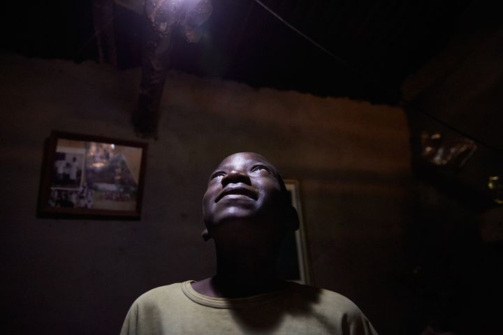 Jan Chumba (12) looks up at one of the light bulbs, Chiuta village, southern Malawi, 2017. Powered by a solar battery, the bulbs offer a safe and cheaper alternative to the more expensive and dangerous paraffin lamp, that Ben and his family used to use. Ben explains “We go to get a battery every 4 days. It is very heavy. We use the light to study and to light the rooms in our house. I like to study. Before the battery we were using paraffin and candles. We like solar much better. Because it provides more light.”