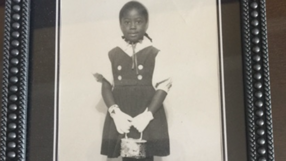 A childhood photo shows a young Trilby Barnes smiling and holding a purse. 