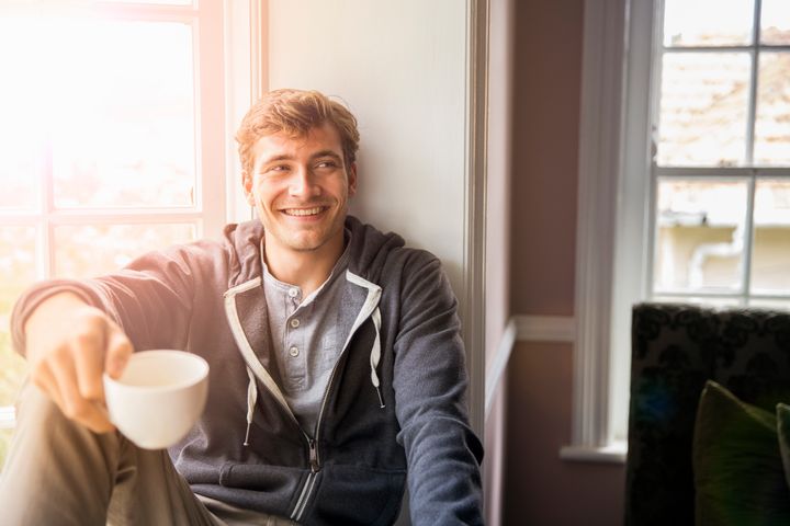 Thoughtful young man holding coffee cup while looking away. Handsome male is wearing hooded shirt by window. He is smiling while sitting at home. Portra via Getty Images