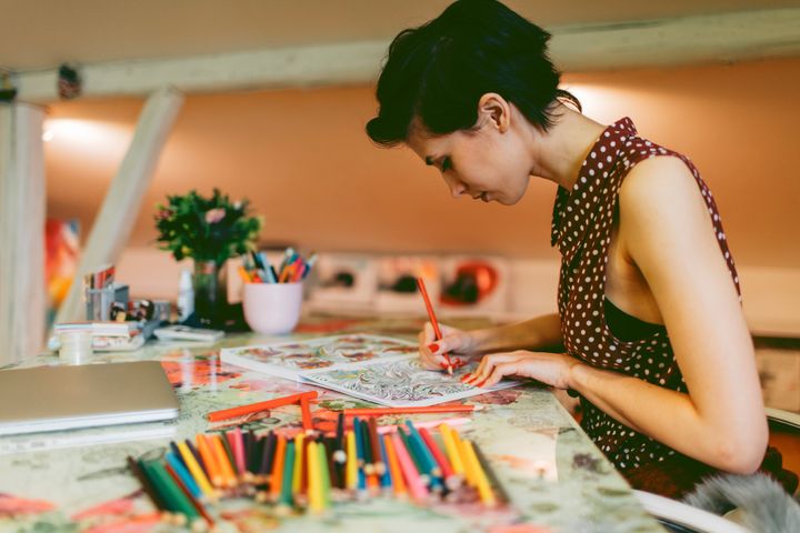 Young smiling woman coloring book for adults in her office or apartment, Enjoy while coloring book for adults. Closed laptop on her desk. vgajic via Getty Images