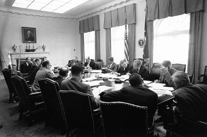 President Kennedy, in the center of the frame writing on a legal pad, presides over an ExComm meeting in the Cabinet Room. To his left is Defense Secretary Robert McNamara; Robert F. Kennedy is slumped in his seat on the far left. With his back to camera wearing glasses is White House Special Counsel and ExComm member Ted Sorensen, later national co-chairman of Gary Hart for President and elder statesman advisor to Barack Obama.
