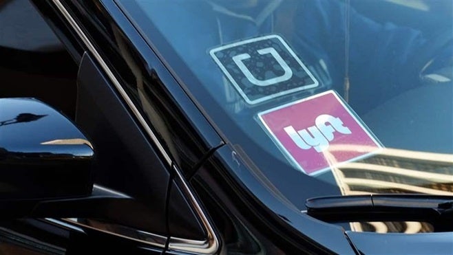 A driver displaying Lyft and Uber stickers on his front windshield drops off a customer in downtown Los Angeles. Some state c