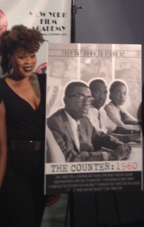 Director Tracy Twinkie Bryd standing alongside the “The Counter: 1960” film poster. 