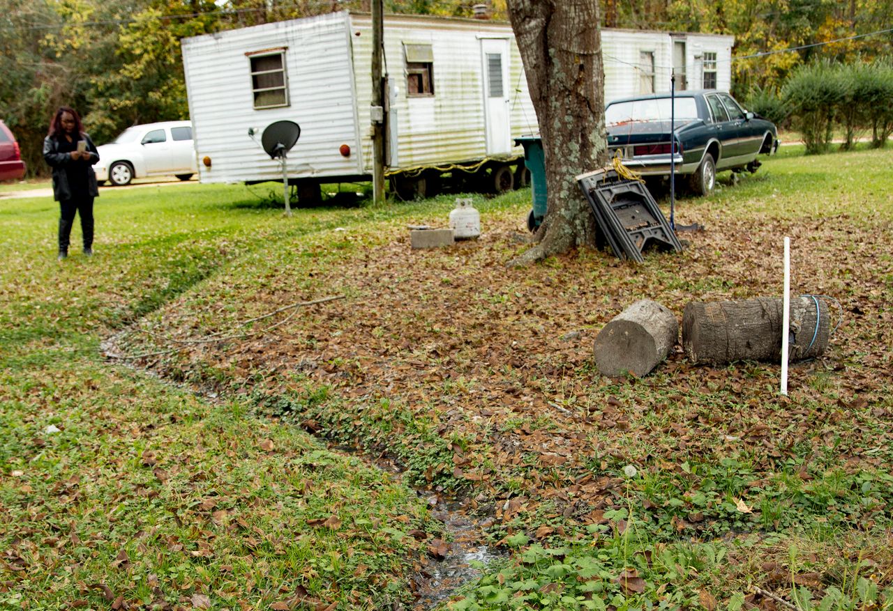 An open sewer carries waste from a mobile home toward the woods at the edge of the property.