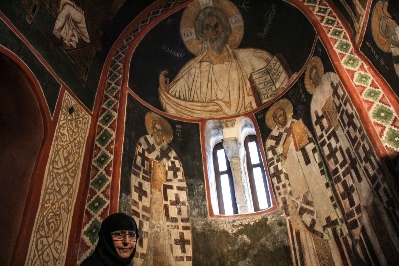Ιερά Μονή Κάτω Παναγιάς: η γερόντισα Χαριτίνη Μπαρμπαρέσου μπροστά από τις τοιχογραφίες (Ο Παλαιός των Ημερών) του 1250 μ.Χ.. Το μοναστήρι παραμένει από τότε αδιάλειπτα ενεργό και οι μοναχές του μοναστηριού, εν έτη 2018, μοιάζουν ο ζωντανός συνδετικός κρικος με ένα μακραίωνο παρελθόν.