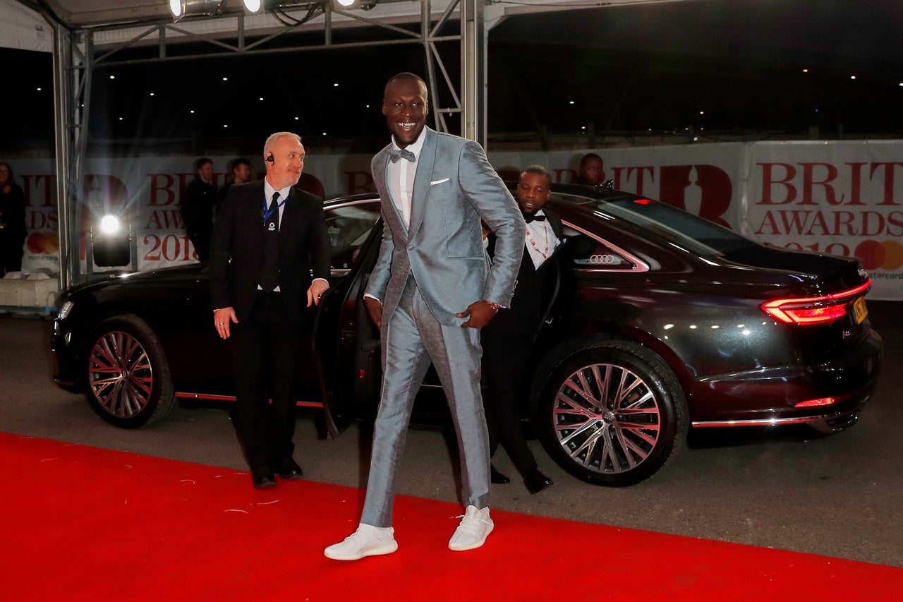 Stormzy arrives at the BRIT Awards wearing a grey suit, bow tie and white trainers.