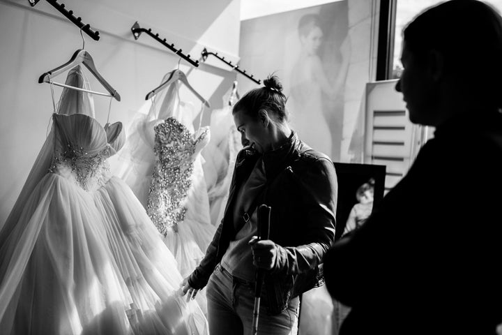 Steph Agnew touches a wedding gown that's been brought out for her to examine while her friend and bridesmaid Jess, who is in shadow, watches and helps guide her.