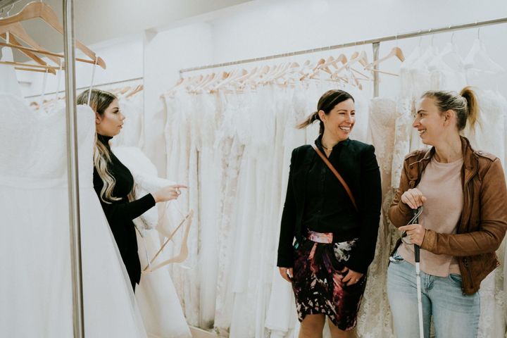 A salesperson, at left, holds a wedding gown in her arms. To the right, Agnew and her friend Jess smile at each other as they're surrounded by racks of white dresses.