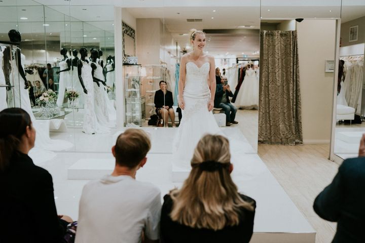 Steph Agnew smiles brightly as she wears a white strapless gown on a small platform in the bridal shop. In the foreground are the backs of her entourage.