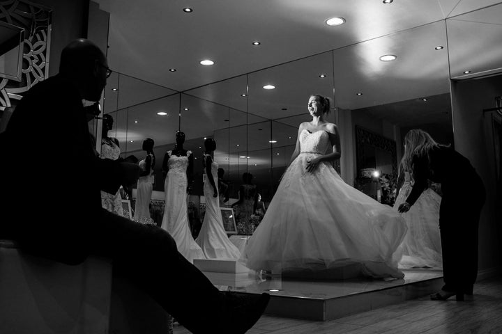 A smiling Steph Agnew stands atop a small platform wearing a flowy, strapless wedding gown. Her dad, who is in shadow on the left, watches his daughter. On the right, a bridal consultant straightens the hem of the gown.