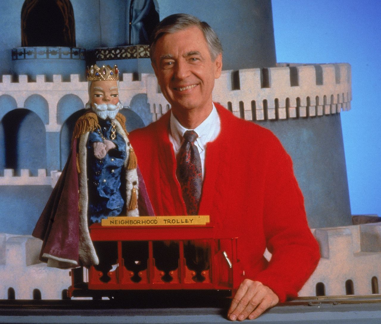 A portrait of Fred Rogers posing with a toy trolley on the set of his television show "Mister Rogers' Neighborhood" in the 1980s.
