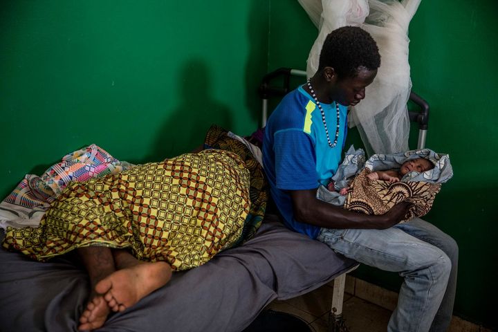 A young man sits with his newborn baby in his lap, on the edge of a bed where the child’s mother is resting, in UNICEF-supported Canchungo Hospital in the Cacheu region of Guinea-Bissau.