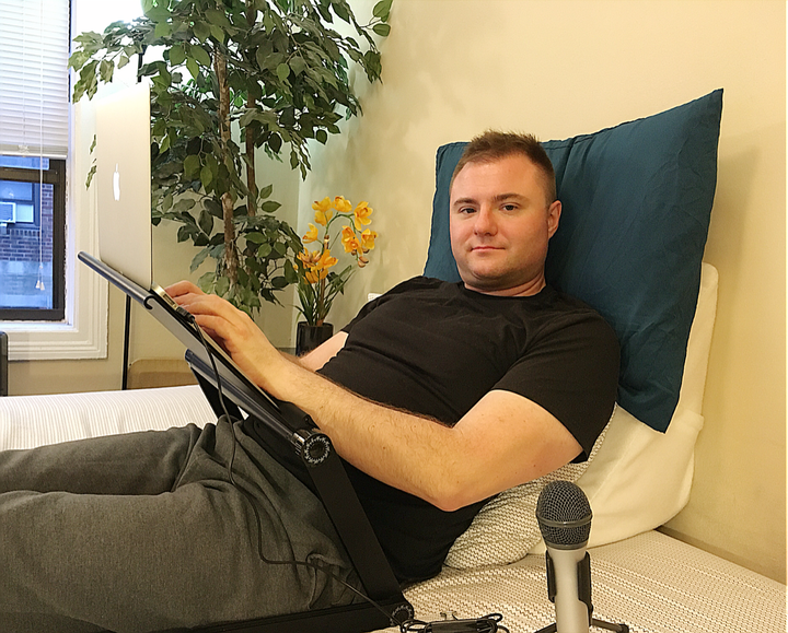 Rick Lunkenheimer lying at a 45-degree angle on a wedge pillow with extra pillow for neck support, configurable desk and microphone for voice dictation.