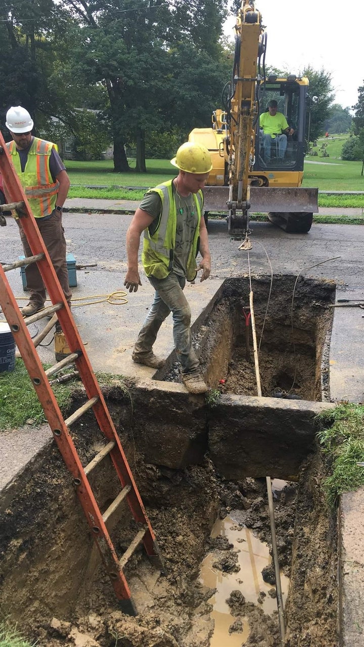 A backhoe pulls old lead piping out while a worker inside the home pushes a new copper pipe through.