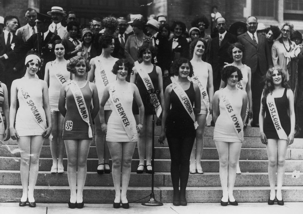 Miss America contestants, representing their home cities and states, pose in one-piece bathing suits and sashes on the steps
