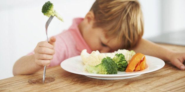 A cute young boy with his head on the table while holding a piece of broccoli on his fork