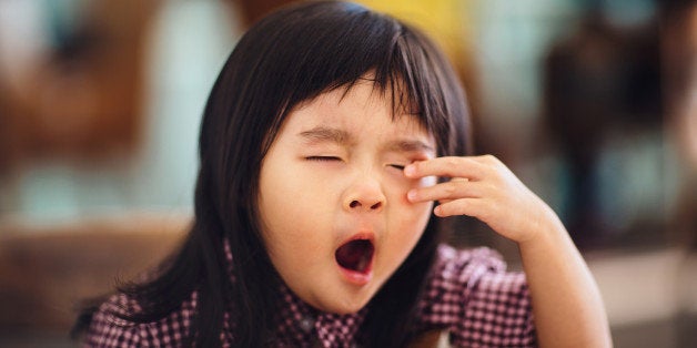 Sleepy toddler girl resting her arm in wooden chair while yawning & rubbing her eyes in a cafe.