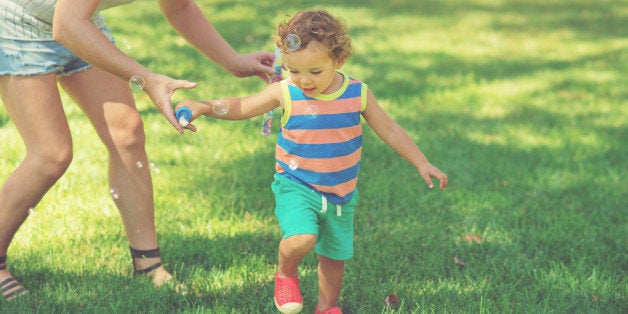 Cute little toddler boy with curly hair getting help from his mother as he plays outdoors with bubbles.