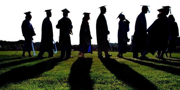 Graduates march onto grassy field during graduation ceremony, sunset and back lighting create silhouette figures.