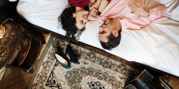 A mature, loving couple is looking at each other lying on the white sheets of a hotel bed. They are holding hands and seem to be in love. On the floor, there's an old carpet, a suitcase and the woman's shoes.