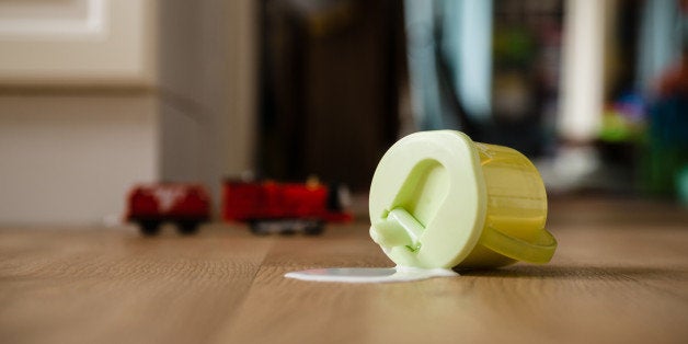 A scene with a tipped over toddler cup and a growing puddle of milk on the floor. The background suggests a domestic setting with a out of focus toy train in the background.