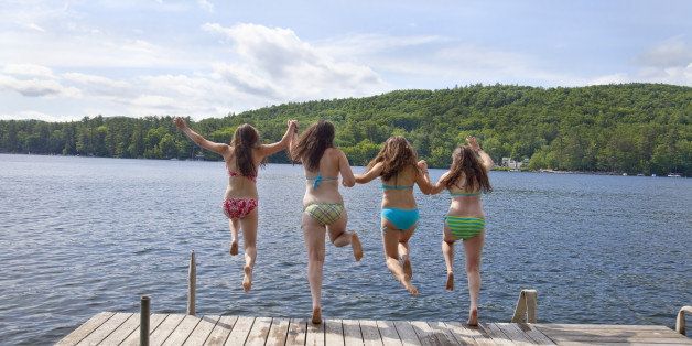 Four teenage girls jumping off a dock at a lake, holding hands. Image depicts the joy of summer vacation and friendship.