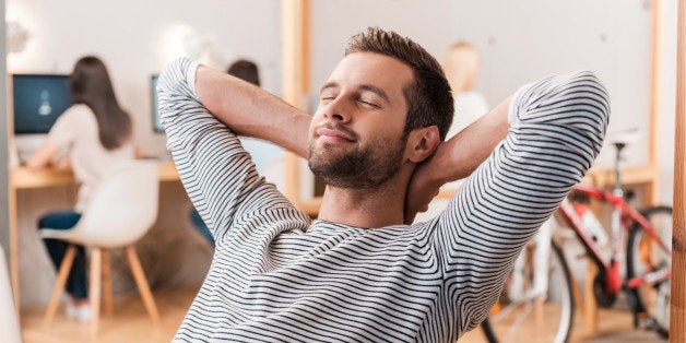 Cheerful young man holding hands behind head and keeping eyes closed while sitting at his working place with his colleagues working in the background