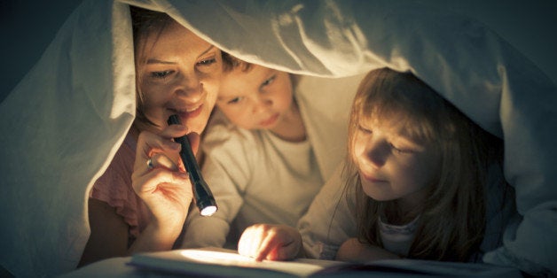 A woman and two children are reading in bed under a sheet. The woman is holding a small black flashlight. The little boy is near the mother. Next to him, the little girl is holding her finger on the page of the book.