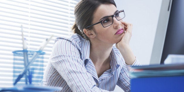 Bored office worker at desk staring at computer screen with hand on chin.