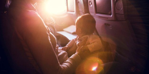 A mother and her baby girl sit in a passenger airline seat, the sun shining brightly in through the plane window. While air travel with children can be difficult, both mom and child are content, the mother with a smile on her face. Horizontal image. INTENTIONAL LENS FLARE.