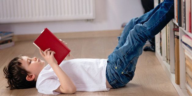 Little boy child reading a book in the library. He lies on the floor. Legs on bookshelf