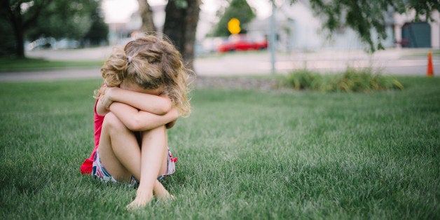 Little girl with curly hair sits on grass and cries into her arms.