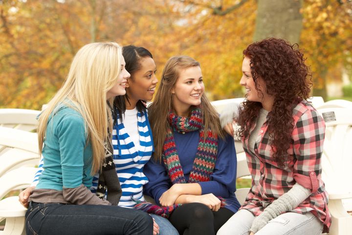 Group Of Four Teenage Girls Sitting And Chatting On Bench In Autumn Park