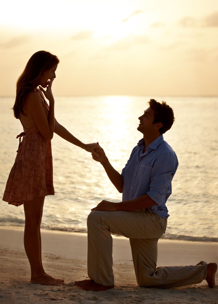 Young man proposing marriage to surprised woman on the beach at sunset