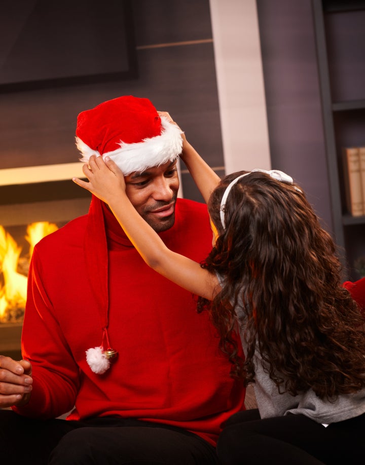 Little girl placing santa hat on father's head at home.