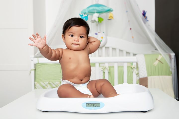 Chubby baby waving hand, sitting on weight scale in nursery room