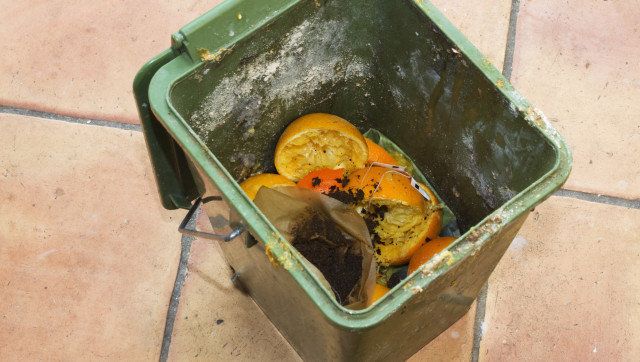 Dirty compost bucket, filled with orange peels and coffee filters, on a kitchen floor.