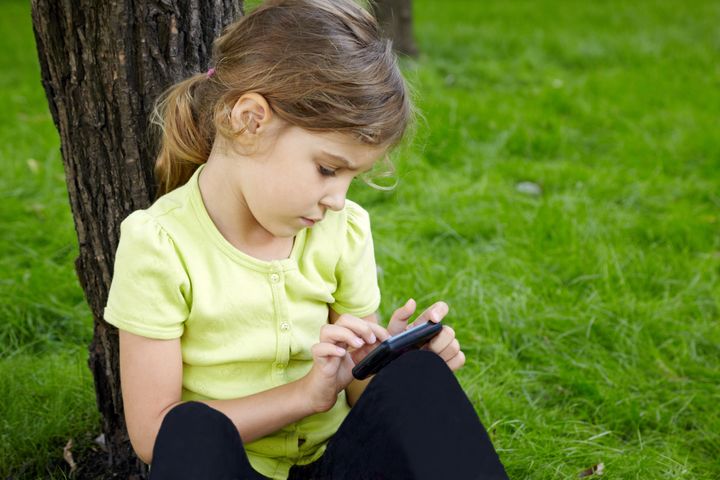Girl sits leaning on trunk of tree in park and plays with cell phone